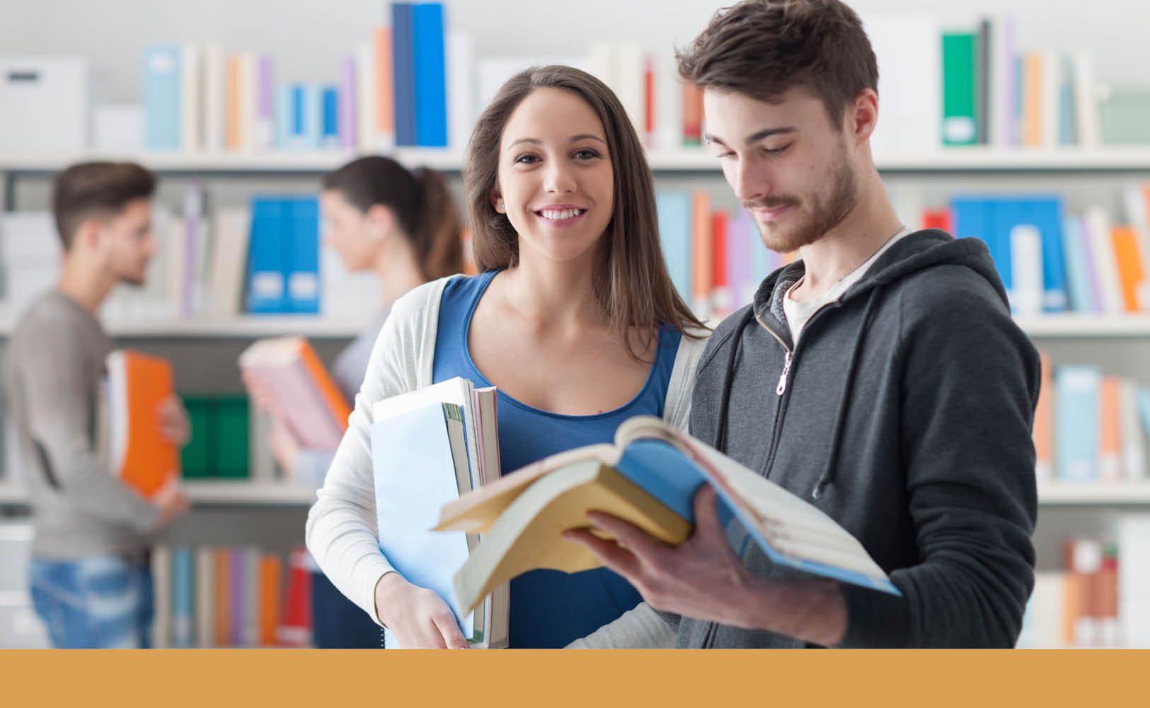 Zwei Studierende in einer Bibliothek: eine lächelnde Frau hält mehrere Bücher, ein Mann blättert in einem aufgeschlagenen Lehrwerk.