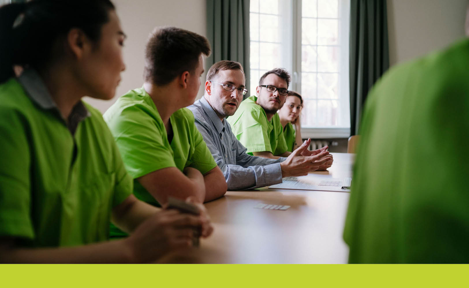 Teilnehmende in grünen Arbeitskitteln sitzen um einen Tisch, ein Mann mit Brille erläutert gestikulierend eine Schulungsunterlage.