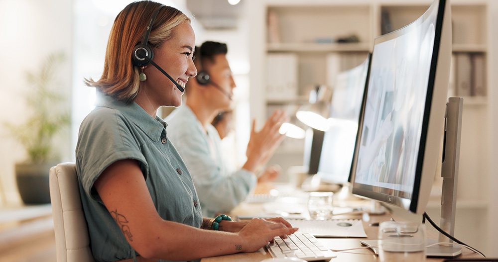 Lächelnde Mitarbeiterin mit Headset am Computer, im Hintergrund weitere Teammitglieder – Symbol für Kontakt und Beratung.